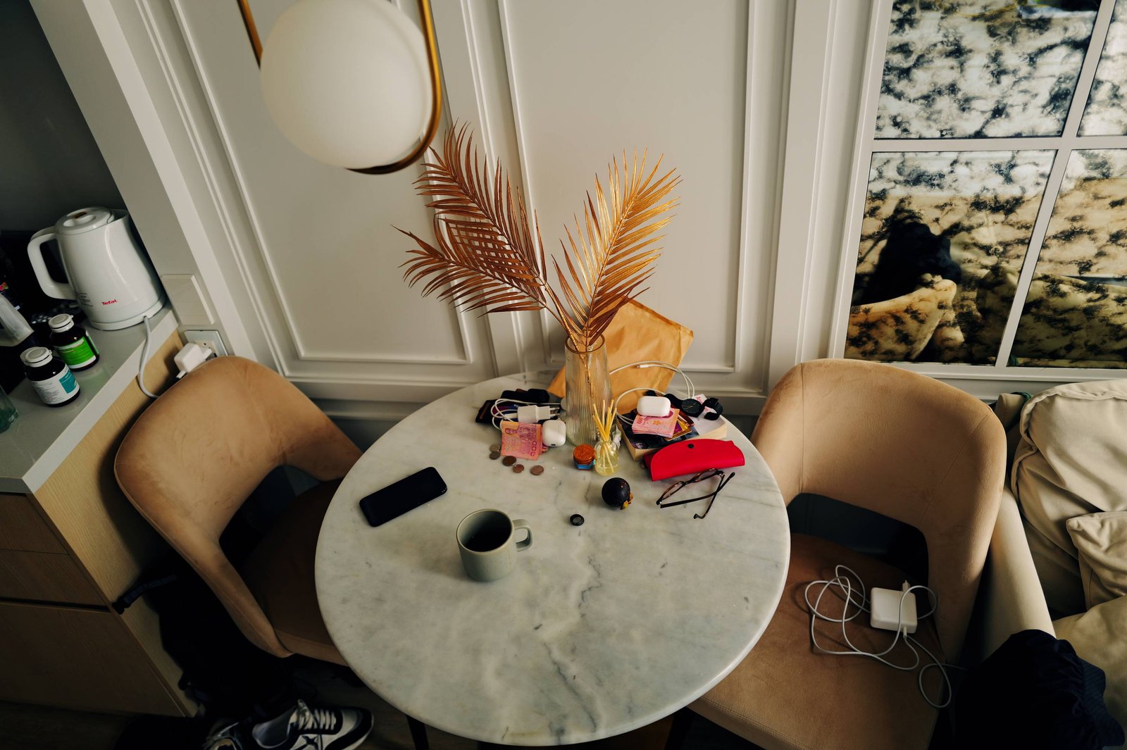 A round marble table seen from above: a coffee cup, a phone, foreign notes and coins, glasses and chargers scattered around gilded palm leaves.