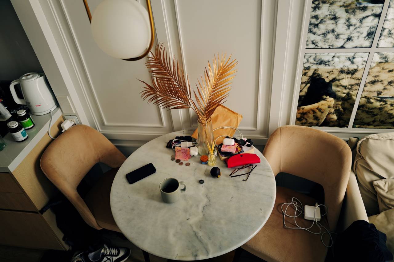 A round marble table seen from above: a coffee cup, a phone, foreign notes and coins, glasses and chargers scattered around gilded palm leaves.