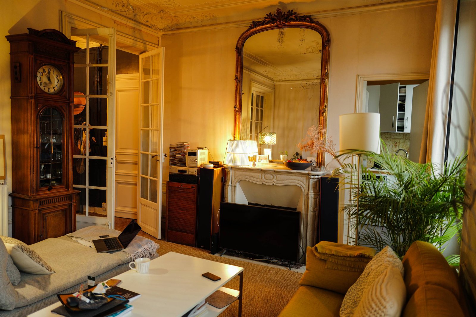 A living room in warm evening light: a grandfather clock, a tall gilt mirror above the marble fireplace, a hi-fi stack, a laptop open on the sofa.