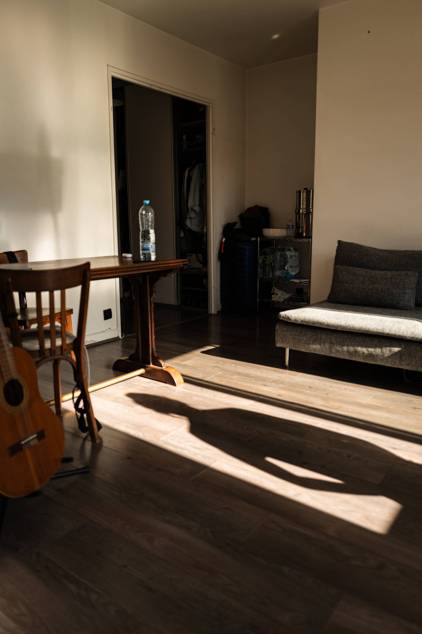 A human shadow stretches across bands of low sunlight on a wooden floor; an acoustic guitar leans in the foreground, a suitcase and shelving by the open doorway.