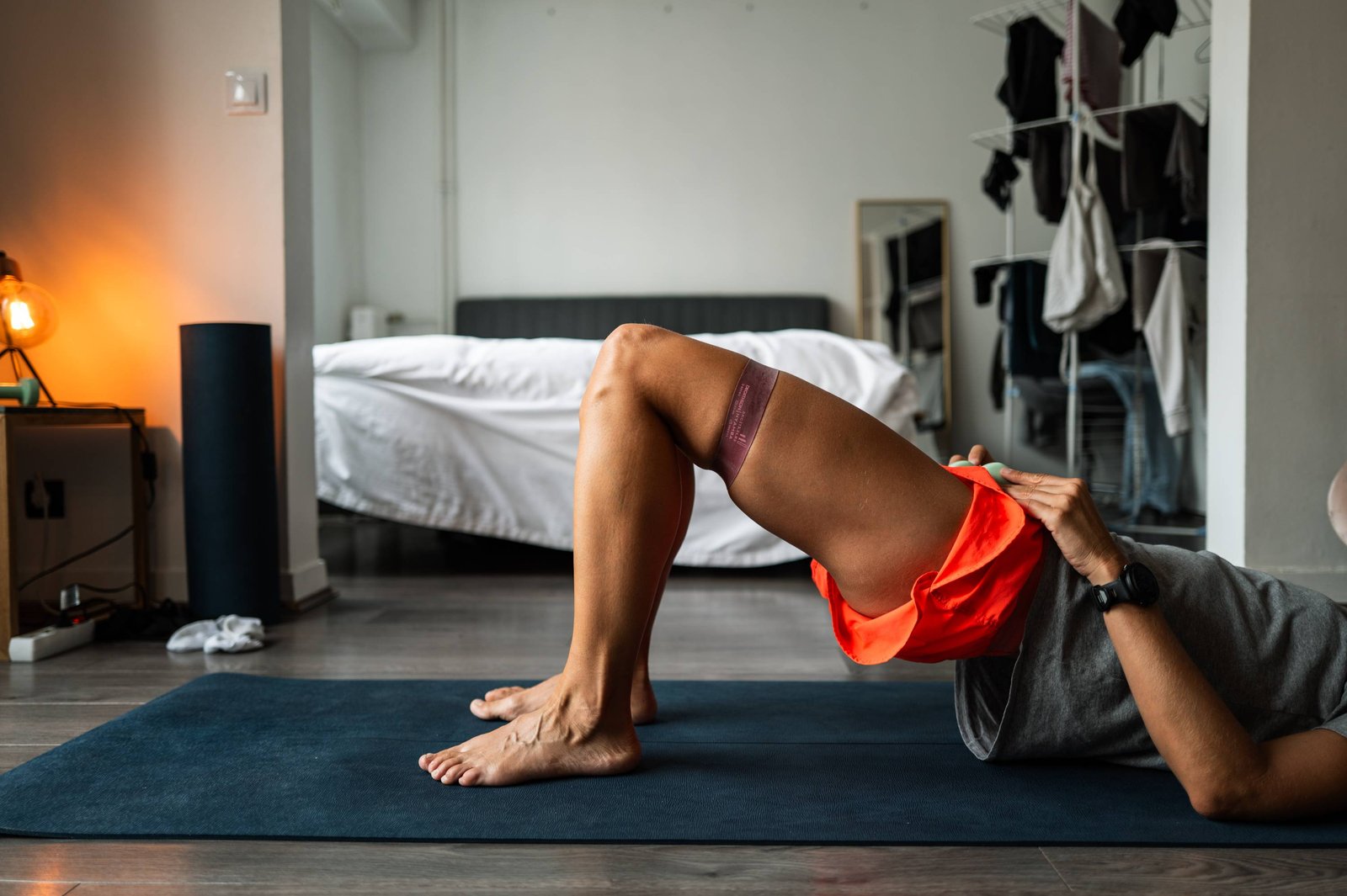A person holds a bridge on a yoga mat in a bedroom, a resistance band around the thighs, the bed and a clothes rail behind.