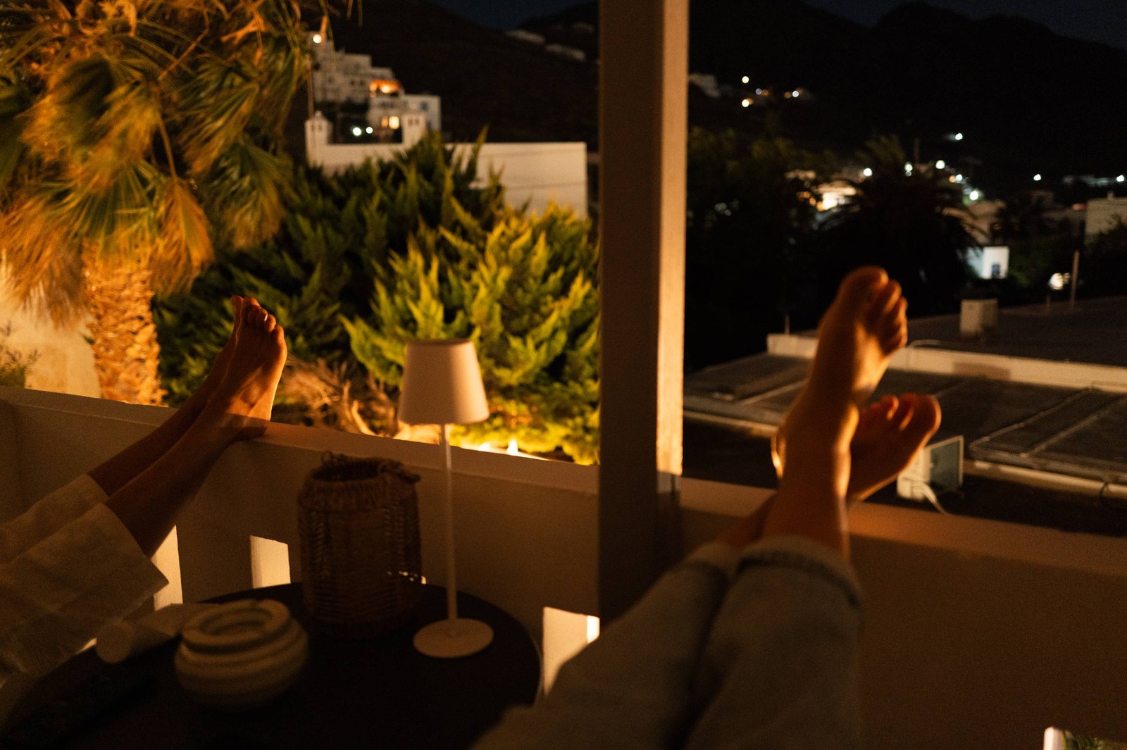 Two pairs of bare feet propped on a balcony ledge at night, a lamp lit among the shrubs, white houses on the dark hillside beyond.