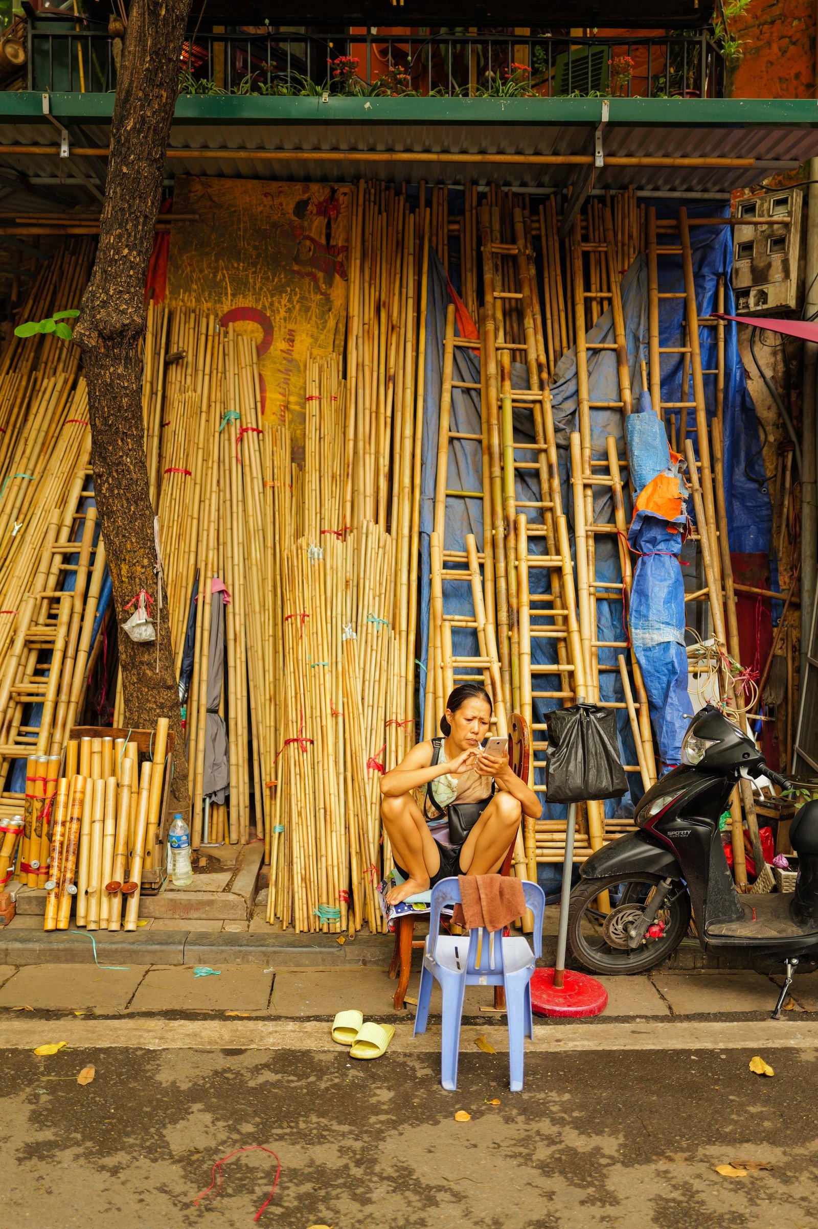 A woman sits barefoot on a low plastic stool in front of a Hanoi shop stacked with bamboo poles and ladders, reading her phone.