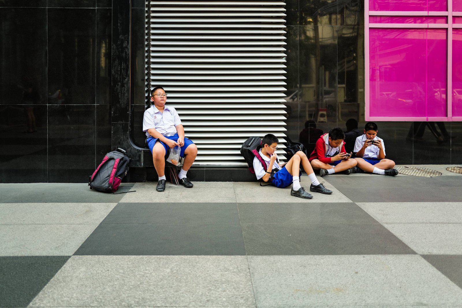 A schoolboy in uniform waits alone on a step against a metal shutter in Bangkok, a bag of snacks in his hands, looking up — no phone.