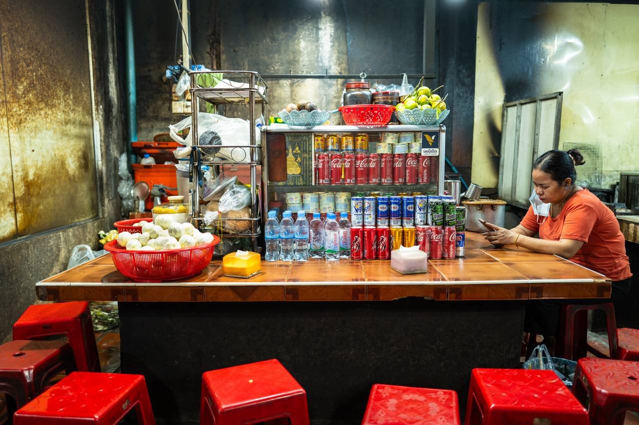 Behind her street food stall in Phnom Penh, a vendor in an orange shirt reads her phone among stacked cans and red stools.