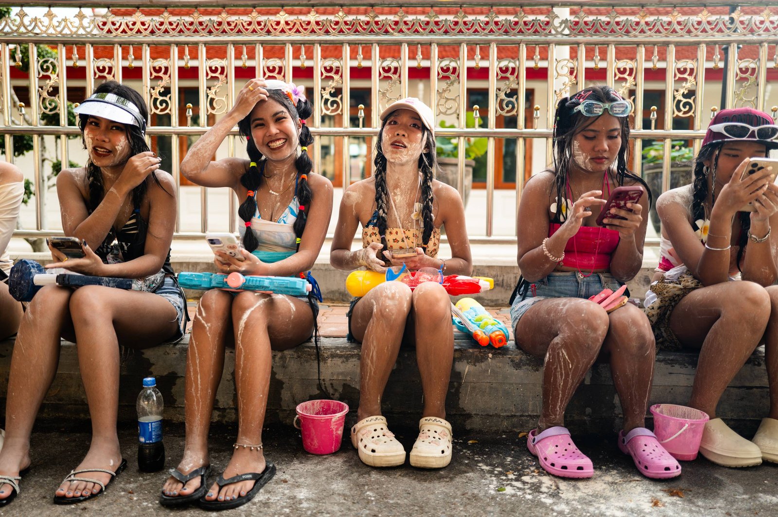 Five young women sit in a row on a low wall during Songkran in Bangkok, faces smeared with white powder and water guns at their feet; two are looking at their phones.