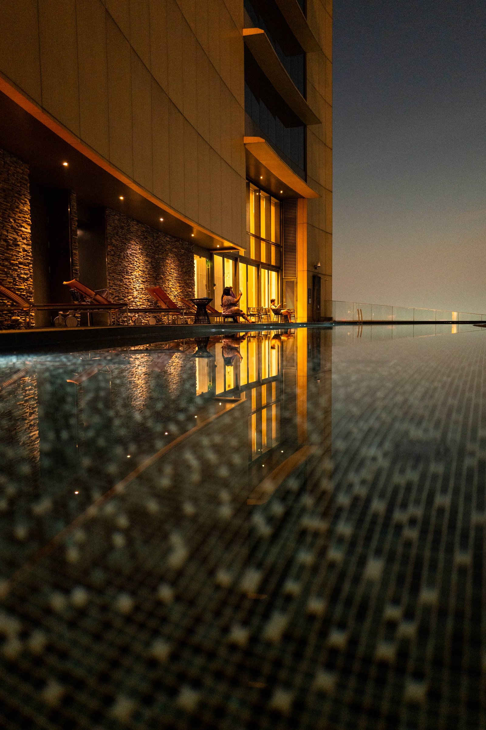 At dusk, a rooftop infinity pool in Bangkok mirrors the lit hotel façade; at the far edge, a figure at the bar is lit by a phone.