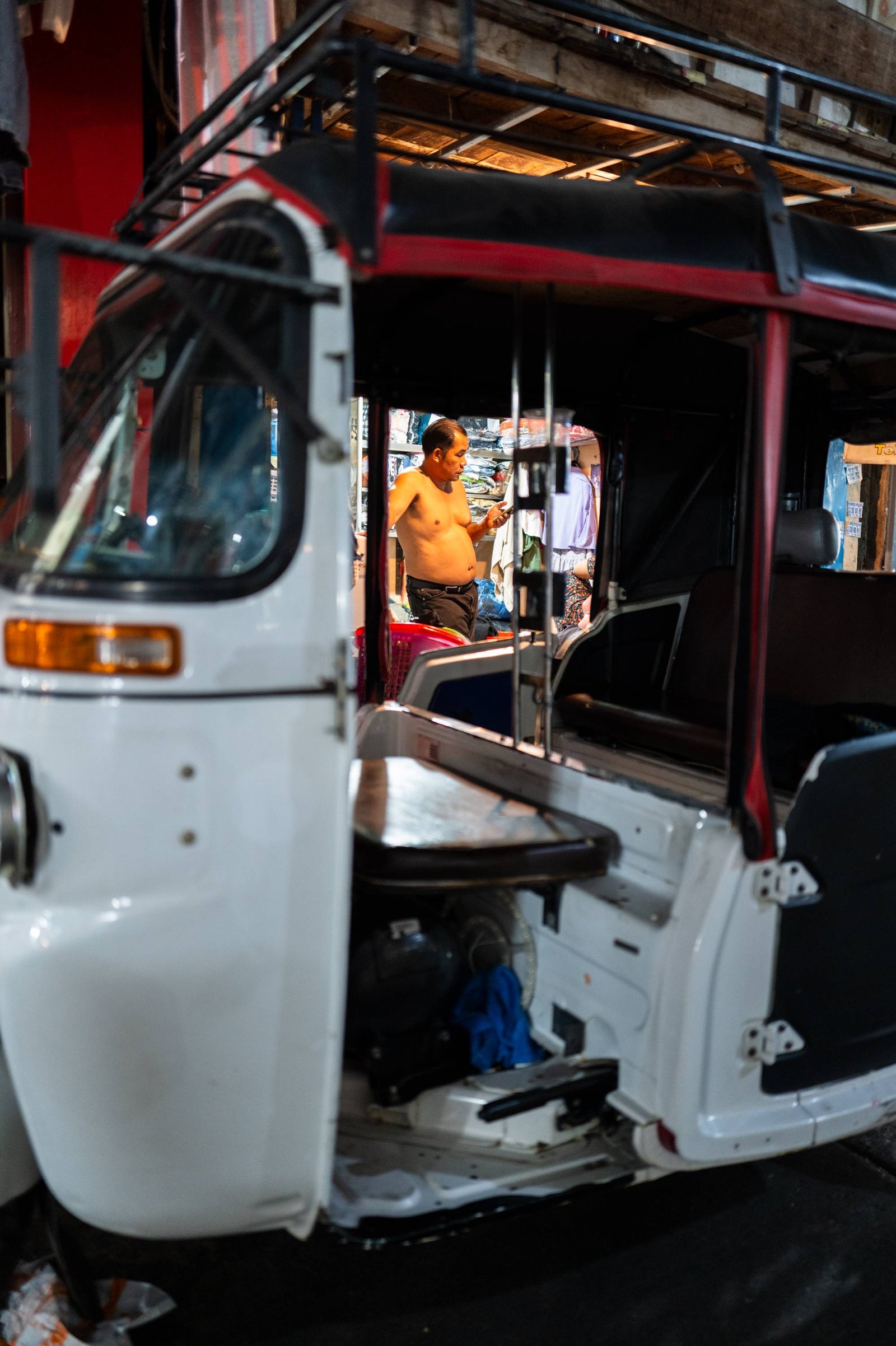 An empty tuk-tuk frames a lit doorway in Phnom Penh, where a shirtless man stands looking down at his phone.