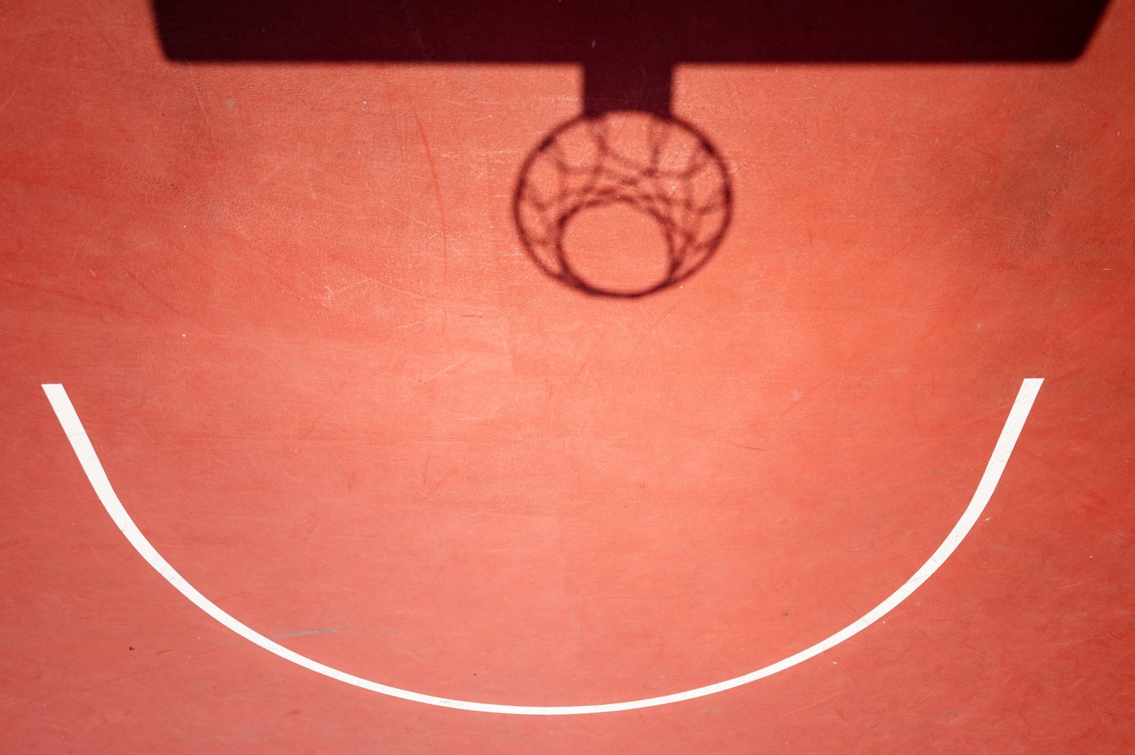 Un terrain de basket vu de face : l'ombre du panier et un arc blanc composent un visage souriant sur le sol terracotta.