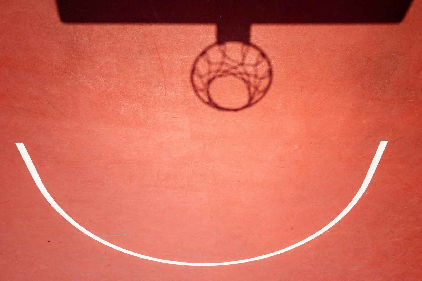 A basketball court seen from the front: the hoop's shadow and a white arc form a smiling face on terracotta ground.