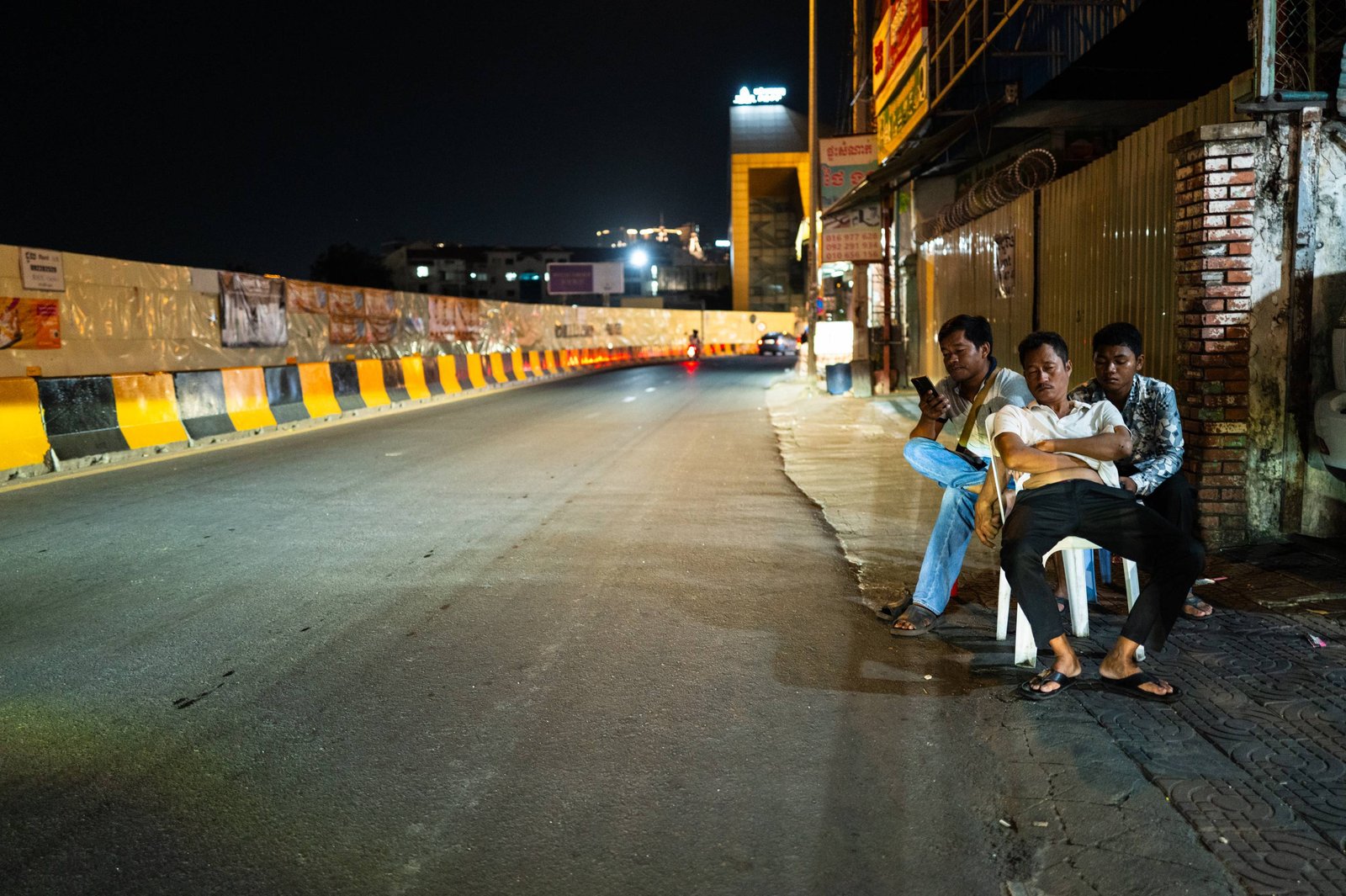 Three men sit on plastic chairs at the edge of an empty road at night in Phnom Penh; one man’s face is lit by his phone screen.