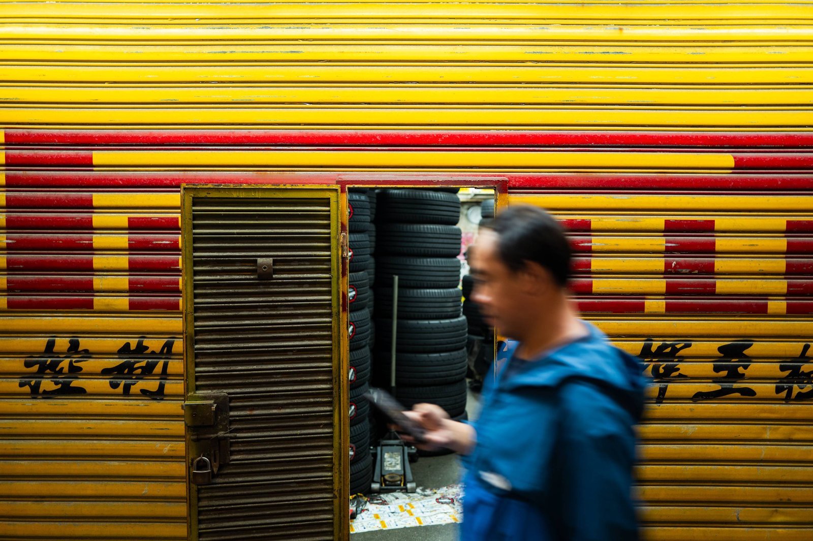 A man in a blue jacket walks past a yellow and red shuttered tyre shop in Hong Kong, phone in hand, blurred by his own movement.