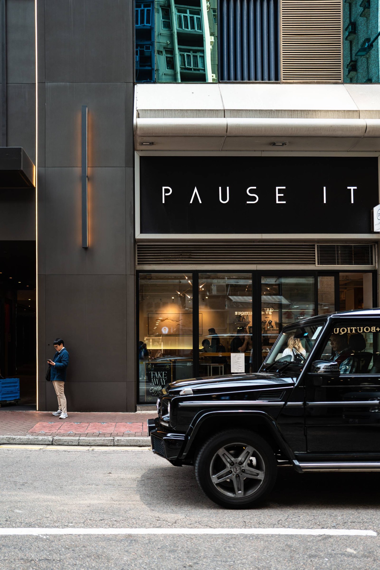 A man checks his phone on the pavement below a Hong Kong shopfront whose sign reads PAUSE IT; a black 4x4 fills the foreground.