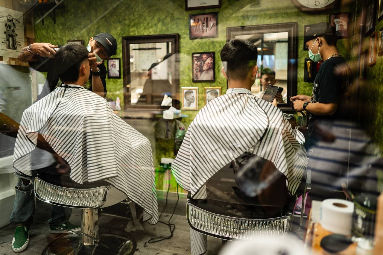 Through a barbershop window, two clients sit under striped capes; one scrolls on his phone while a masked barber cuts the other’s hair.