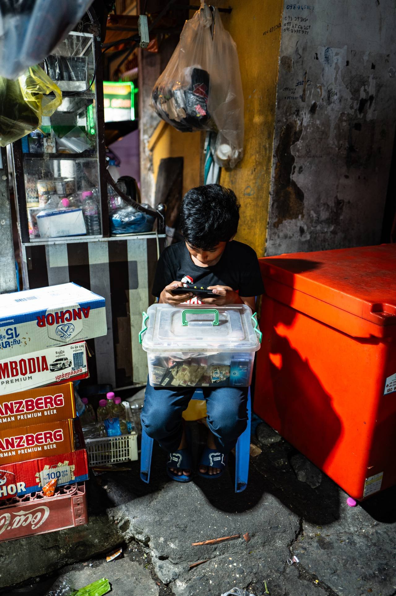 A boy sits on a low stool in an alley, a clear box of small goods on his knees, absorbed in his phone between stacked beer crates and a red cooler.